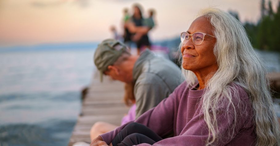 Side view of a beautiful and vibrant senior woman of Pacific Islander descent sitting on a dock with her multigenerational family and watching the sun set with a peaceful expression on her face on a warm summer evening in the Pacific Northwest region of the United States.