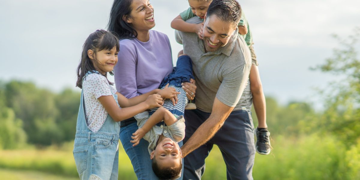 A small family of five are seen playfully enjoying each other company while outside on a sunny summers evening.  They are each dressed casually and are smiling.